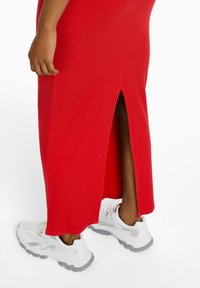 Robe maxi rouge côtelée avec une fente sur le côté. Associée à des chaussures de sport blanches avec des accents gris et une semelle texturée.