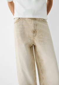 Beige vida jeans, tillverkade av denim, med en urblekt textur och subtila färgvariationer samt standardfickdetaljer.