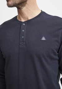 Marineblå langermet henley-skjorte laget av teksturert stoff, med tre knapper ved halsen og et trekantet logo-lapp på brystet.