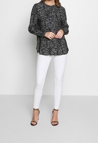 Haut noir à manches longues avec motifs abstraits blancs, associé à un jean skinny blanc et des talons noirs à brides. Tissu lisse avec une coupe décontractée.