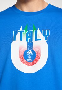 Zila krekls ar uzrakstu "Italy" un baltu U formas elementu ar zaļo un sarkano gradientu, Adidas logo un UEFA Sieviešu Eiropas čempionāta Šveice 2025 emblēmu.