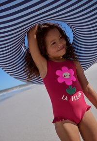 Rode badpak met een roze bloemmotief en ruches, gedragen door een gestreepte opblaasbare ring, tegen een strandachtergrond.