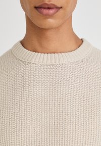 Beige geribbelde trui met een textuur gebreide stof, met een ronde, geribbelde halslijn en een gladde stof. Close-up van de kraag en het bovenste deel van de borst.