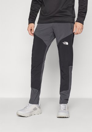 Homme portant un pantalon noir et gris The North Face et des chaussures de randonnée blanches, debout devant un fond uni.