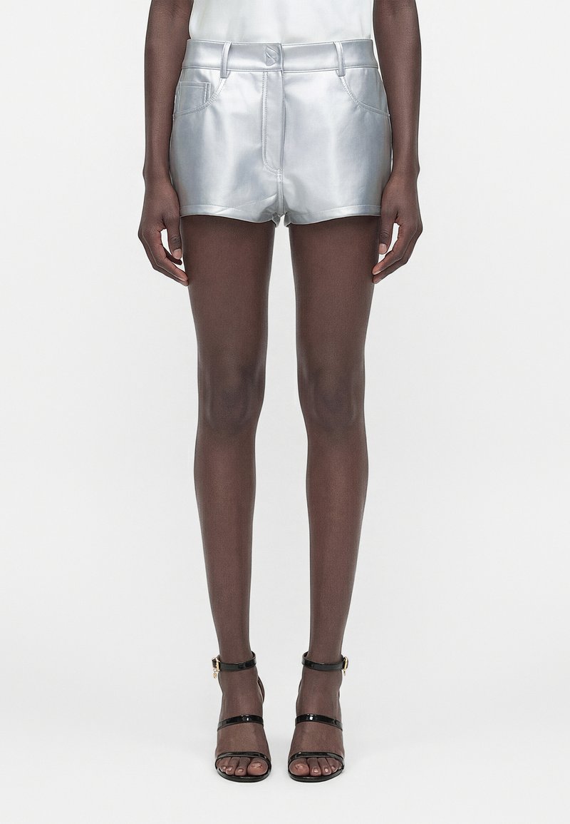 Personne portant un short métallique argenté avec des poches avant et des sandales noires à talons hauts avec brides, se tenant devant un fond uni.