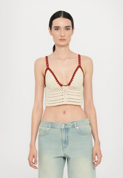 Crochet-style crop top in cream with red accents, featuring a plunging neckline, wide straps, and textured patterns. Paired with light-blue jeans.