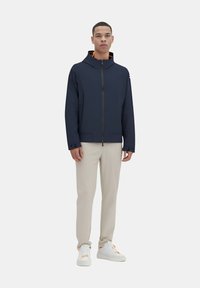 Giacca a vento blu navy con cappuccio e zip full zip, due tasche laterali e interno arancione a contrasto. Indossata con pantaloni color crema e sneaker bianche.