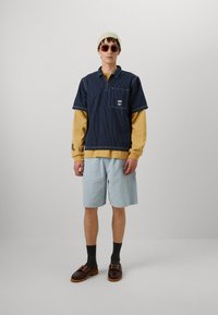 Camicia a righe navy a maniche corte sopra una maglietta a maniche lunghe color senape, pantaloni corti azzurri con texture, scarpe marroni, calzini grigi e un berretto beige.