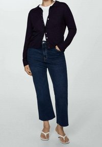 Námořnický svetr s knoflíkovým zapínáním, kombinovaný s bílým tričkem a tmavými džíny. Model nosí bílé žabky a stojí na světlém pozadí.