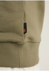 Sudadera verde oliva con dobladillo acanalado y panel lateral, que presenta una etiqueta de tela negra y marrón en la parte inferior izquierda. Material de algodón suave.