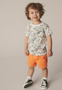 T-shirt grafico bianca con design colorati abbinata a pantaloni corti arancioni. Sneakers nere con lacci bianchi completano l'outfit.