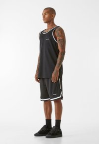 Melna sporta tank top un šortu komplekts ar baltām akcentiem, brīvs piegriezums, apaļa kakla izgriezums un logo detaļas. Valkāts ar melniem sporta apaviem un zeķēm.