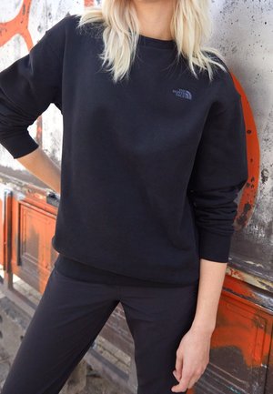 Personne aux cheveux blonds mi-longs portant un sweat-shirt noir The North Face et un pantalon noir, debout contre un mur usé avec des marques orange.