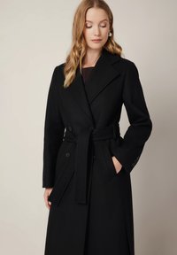 Czarny wełniany trench coat o podwójnym zapięciu, szerokich klapach, pasku i dwóch bocznych kieszeniach; gładka faktura z dopasowanym krojem.