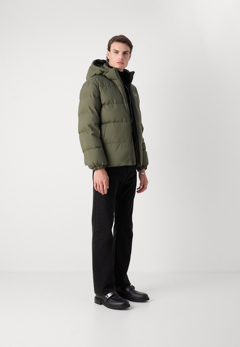 Olivgrüne Steppjacke mit Kapuze, elastischen Bündchen und Reißverschluss, kombiniert mit schwarzen Jeans im Straight-Leg-Schnitt und schwarzen Schuhen.