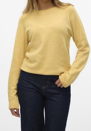 Kvinde iført en blød gul langærmet sweater, der er let stoppet ind i mørkeblå højtaljede jeans, stående med den ene hånd i lommen.
