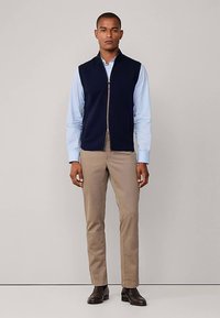 Veste sans manches bleu marine avec une fermeture éclair à l'avant, portée sur une chemise à manches longues bleu clair, associée à un pantalon beige et des chaussures foncées.