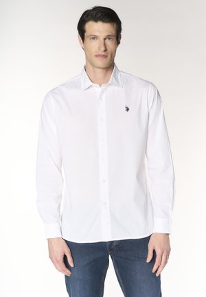 Uomo che indossa una camicia bianca a maniche lunghe con un piccolo logo scuro ricamato sul petto e jeans blu.