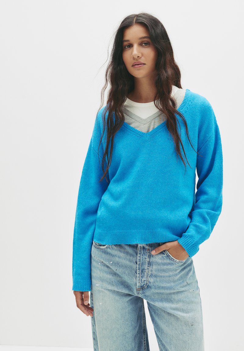 Blauer Strickpullover mit V-Ausschnitt, langen Ärmeln und geripptem Saum, kombiniert mit hellblauen Jeans mit Farbspritzern.