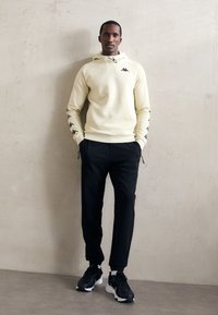 Man standing against a light wall wearing a cream hoodie with black logos on sleeves and chest, black pants, and black sneakers with white soles.