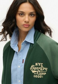 Femme avec des cheveux sombres et ondulés portant une chemise boutonnière bleu clair sous une veste vert foncé avec "NYC SuperDry Corp" brodé sur la poitrine.