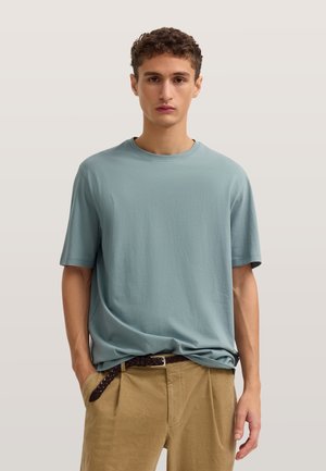 Junger Mann trägt ein hellblaues kurzärmeliges T-Shirt, das leicht in beige Hose mit braunem geflochtenem Gürtel gesteckt ist, steht vor neutralem Hintergrund.