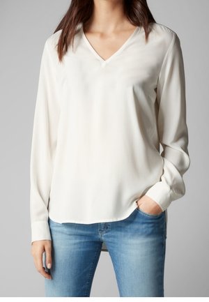 Kvinde iført en langærmet hvid V-hals bluse kombineret med blå jeans, den ene hånd i lommen, foran en ensfarvet grå baggrund.