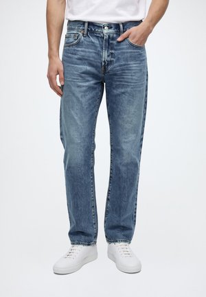 Personne portant un jean bleu droit et des baskets blanches, debout avec la main droite dans la poche sur un fond clair uni.
