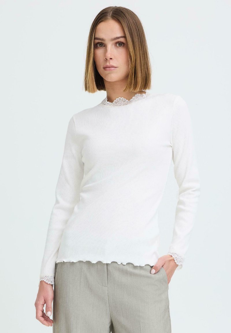 Femme aux cheveux raides mi-longs portant un haut blanc à manches longues avec bordure en dentelle et un pantalon gris clair, debout devant un fond uni.