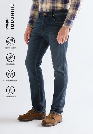 Man draagt donkerblauwe jeans, bruine schoenen en een geruit overhemd, met tekst en iconen die het lichtere gewicht, de stevigheid, technisch voortgestuurde spijkerbroek en beweging benadrukken.