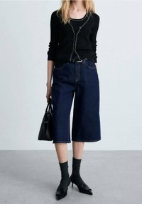 Haut à manches longues noir côtelé, culottes en denim larges bleu foncé, chaussettes noires à la cheville, chaussures à talons noires et sac à main noir.