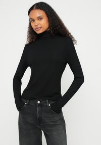 Bluza neagră cu mâneci lungi și guler înalt, realizată dintr-un material moale, cu un design ajustat și textură netedă, asortată cu blugi închiși la culoare.