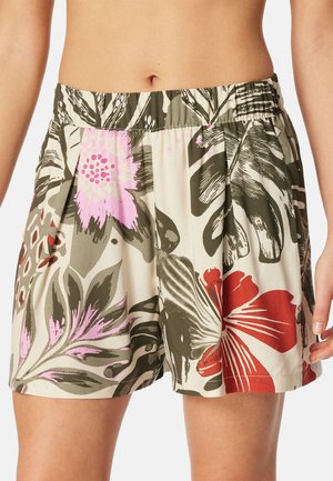 Žena nosí volné šortky s elastickým pasem, na béžovém pozadí s velkým květinovým vzorem v růžové, červené a zelené barvě.