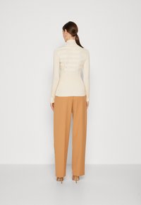 Beige strukturierter Oberteil mit hohem Kragen und gestreiftem Muster, kombiniert mit hochtaillierten, weit geschnittenen, hellbraunen Hosen und nude-farbenen Absätzen.