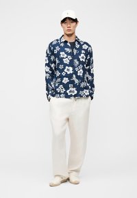 Νεαρός άνδρας φορώντας μπλε σκούρο πουκάμισο με λουλουδάτο και λεοπάρ σχέδιο, λευκό παντελόνι με φαρδιά μπατζάκια, λευκά slip-on παπούτσια και λευκό καπέλο, στέκεται μπροστά σε λευκό φόντο.