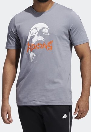 Homme portant un t-shirt gris avec un crâne blanc et un texte rouge stylisé, associé à un pantalon noir Adidas.