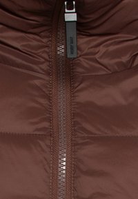 Brown puffy vest with a central zipper, featuring a nylon exterior and horizontal stitched panels for insulation. Zipper pull has a branded tag.
