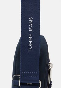 Bolsa de tela azul marino con un acabado texturizado, con un zip plateado visible y una correa etiquetada con "TOMMY JEANS" en letras blancas.