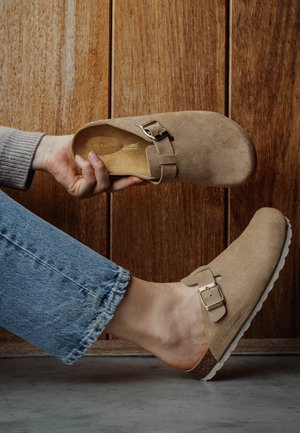 Zuecos de ante en beige claro con hebilla ajustable, plantilla de corcho y suela de goma blanca. Textura suave con un diseño minimalista.