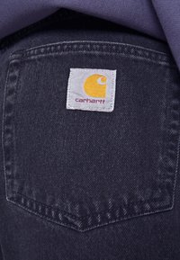 Takapokka tummansinistä farkkua olevissa housuissa, joissa on valkoinen Carhartt-tunnus, jossa on keltainen logo ja punainen teksti.