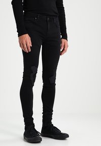 Jeans negros skinny con detalles de rodillas rasgadas, fabricados en denim elástico, con una cintura baja y estilo de cinco bolsillos, combinados con zapatillas negras.