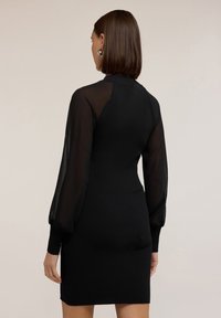 Schwarzes geripptes Kleid mit langen transparenten Puffärmeln, taillierter Silhouette und hohem Ausschnitt. Der Stoff hat eine glatte Textur mit einem subtilen Glanz.