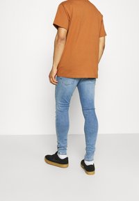 Camiseta de algodón marrón, vaqueros ajustados azules, zapatillas negras con suelas de goma, mostrando un look casual desde atrás. Costuras mínimas y sin logotipos visibles.