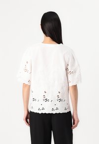 White embroidered top with short sleeves, featuring cut-out floral patterns and a scalloped hem. Back view with simple round neckline.