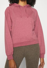 Sweat-shirt rose avec capuche, manches longues raglan, poignets côtelés et ourlet ajusté. Fabriqué en tissu doux et texturé. Associé à des leggings marron.