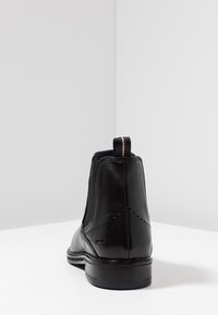 Botas de cuero negras hasta el tobillo con paneles laterales elásticos, puntera redondeada, perforaciones decorativas y una tira de agarre a rayas en la parte trasera.
