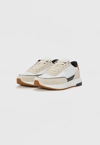 Adidași bej și albi din materiale din suede și plasă, cu un accent negru și talpă din cauciuc texturat. Design cu șireturi și branding minim.