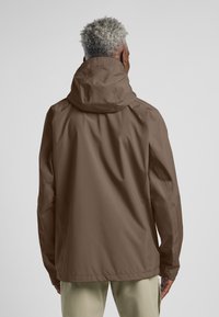 Giacca impermeabile marrone con cappuccio, caratterizzata da una texture liscia e un design minimalista. La parte posteriore mostra dettagli delle cuciture e un fit rilassato.