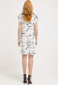 Abito bianco con motivo a linee nere astratte, maniche corte e dettagli decorativi sulle spalle. Indossato con scarpe con tacco alto nere.