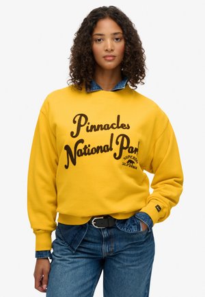 Gul sweatshirt med sort skrift "Pinnacles National Park" og logo. Ribbet manchetter og kant, parret med denimjeans, let tekstur.